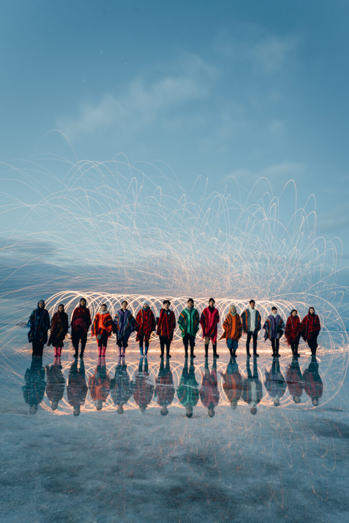 鏡張りのウユニ塩湖で光の軌跡を背景に立つカラフルなポンチョ姿の参加者